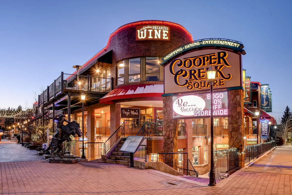 The vibrant exterior of Cooper Creek Square, with its inviting shops, dining options, and artistic statue. The scene is lively, with people gathering, shopping, and enjoying entertainment in a festive, welcoming setting.