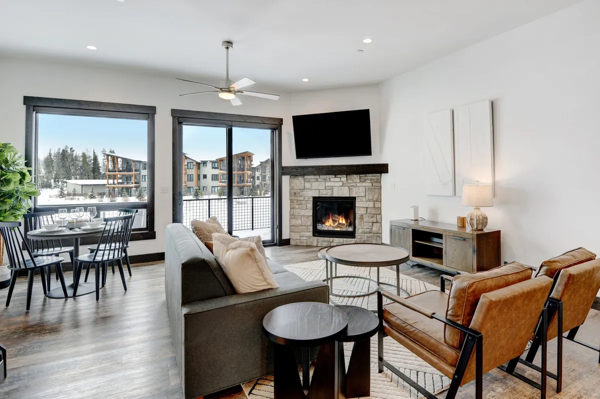 A spacious, light-filled living area with a roaring fireplace, large windows framing snowy outdoor scenery, a dining table ready for gatherings, and a cozy seating arrangement. The setting evokes warmth, comfort, and a place to unwind after an active day.