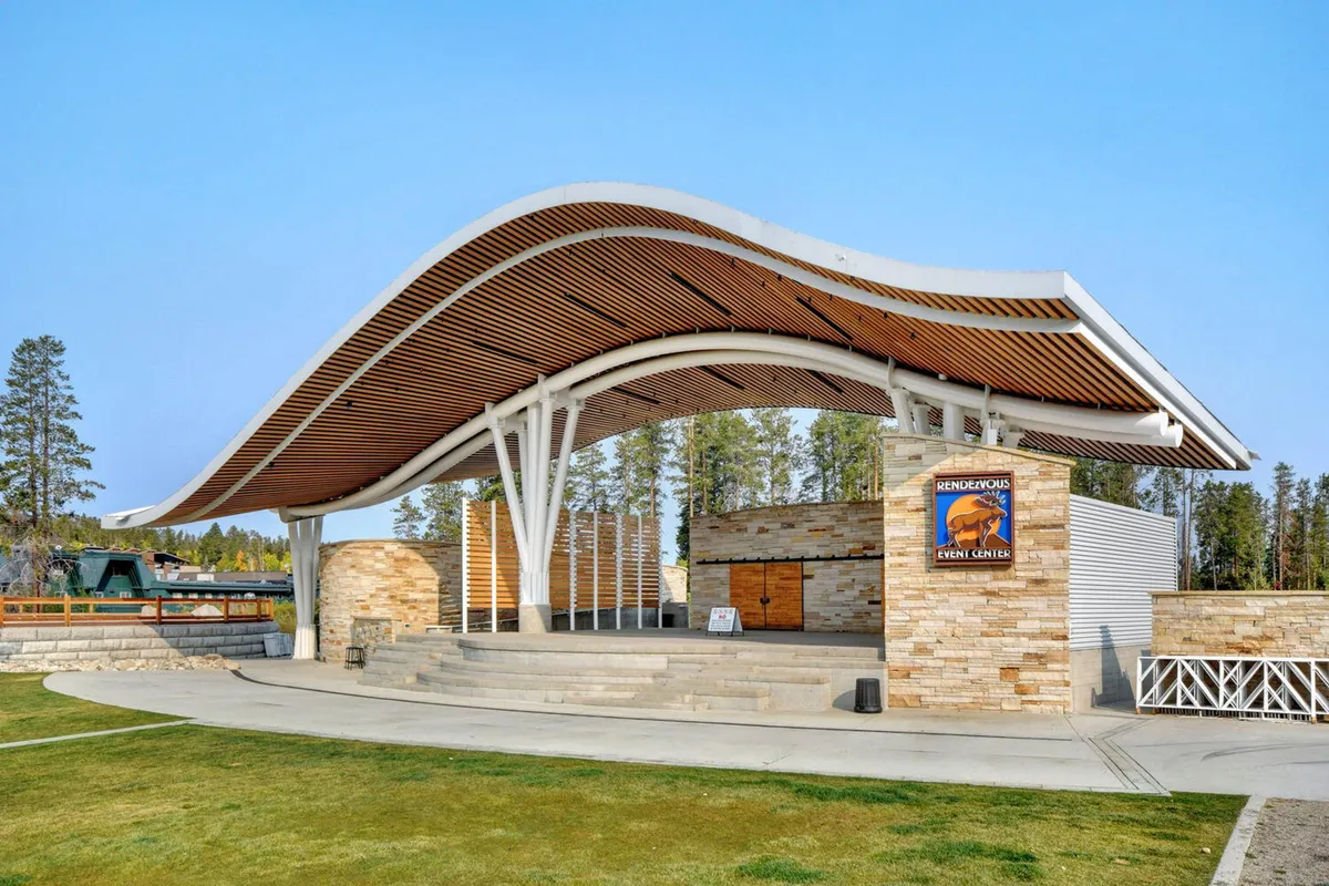 A lively outdoor amphitheater with a curved roof structure, stone facade, and prominent event signage. The space is bustling with families and community members attending concerts or local events, capturing the celebratory and social atmosphere of the neighborhood.