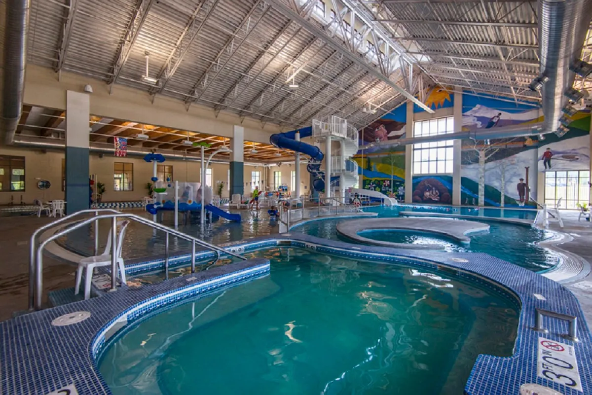 A vibrant interior scene of a recreation center pool area, complete with a waterslide, colorful murals, and children playing. Lounge chairs invite relaxation and the atmosphere is lively and family-friendly.
