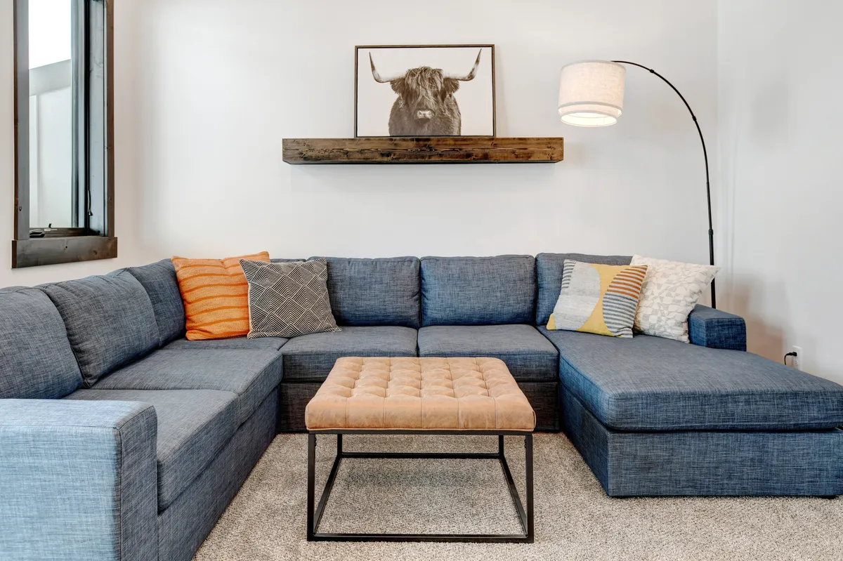 A cozy interior space featuring a large blue sectional sofa adorned with orange and grey throw pillows. There's a modern coffee table in the center, a floor lamp for ambient lighting, and eye-catching bull artwork on the wall. The setup invites relaxation, socializing, or family activities.