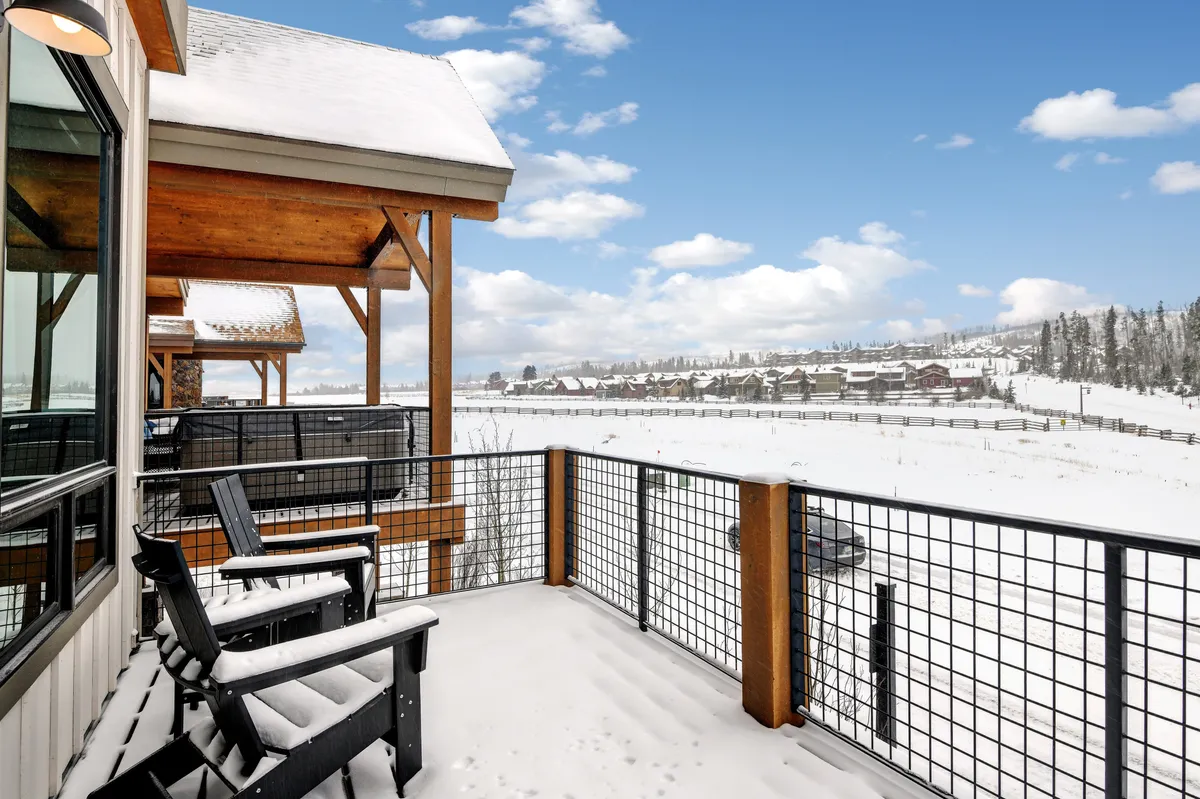 This exterior image displays a luxurious balcony overlooking a serene, snow-covered mountain landscape, with distant houses and a clear blue sky in the background. People are relaxing on the balcony, enjoying the winter scenery and each other's company.