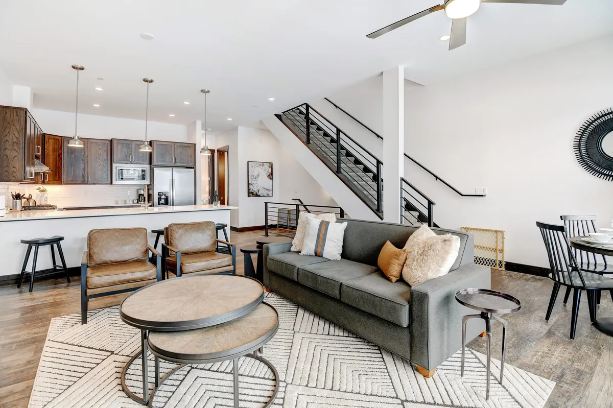 An open-plan space seamlessly connecting the living room, dining area, and kitchen. The view includes a cushioned sofa, a coffee table, a kitchen island with barstools, a staircase, and a dining table, showing the effortless flow between gathering spaces.