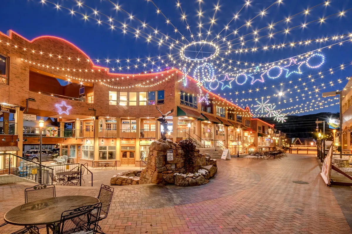 A cheerful exterior scene featuring string lights, a central fountain, decorative elements, and surrounding buildings, where people are strolling, dining, and socializing. The atmosphere suggests an inviting local hub, ideal for exploring breweries, shops, or live entertainment.