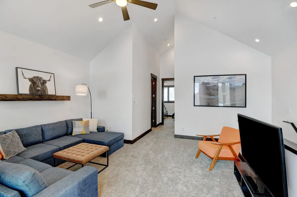 An inviting living area with a comfortable sectional sofa, a bright orange accent chair, modern wall art, and a flat-screen television. The arrangement is open and relaxed, ideal for lounging, watching TV, or social time.
