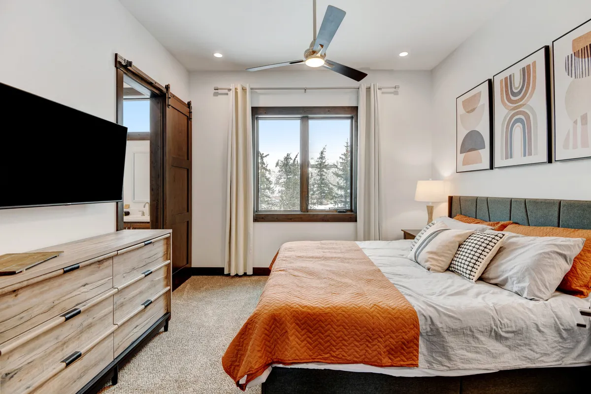 A serene bedroom scene showcasing a bed with a cozy orange blanket, a large window with a scenic view, wall art, and a TV atop a dresser. The space is perfect for relaxing, reading, or enjoying a movie in bed.