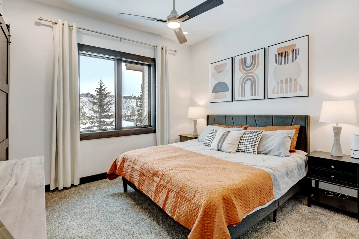 A beautifully appointed bedroom featuring a bed with an orange quilt, a window with tranquil mountain views, and decorative artwork. The inviting linens and serene color palette evoke comfort and relaxation.