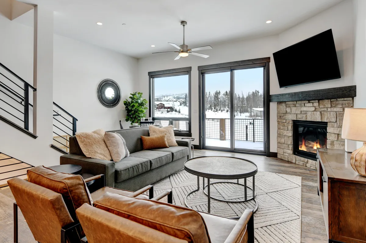 A spacious living room filled with natural light from large windows offering snowy mountain views. The room features a striking custom stone fireplace, a comfortable sofa, stylish armchairs, a decorative mirror, and cozy accents that create a warm, inviting setting for relaxation and socializing.